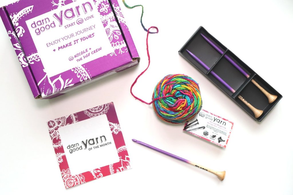 Sigoni is showing all of the items included in the Budget-Friendly Crochet Subscription Box from Darn Good Yarn of the Month. The subscription box includes: welcome booklet, 100% recycled silk yarn (rainbow colored), 5mm wooden crochet hook, 5mm wooden knitting needles
