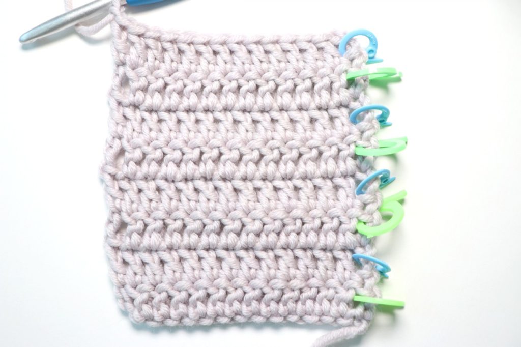 In this photo, Sigoni Macaroni is explaining how you should be counting your crochet stitches and rows using a swatch of double crochet. Pictured is a yarn swatch consisting of eight rows of double crochet stitches. There are stitch markers showing the alternate rows and arrows to mark where each row begins.